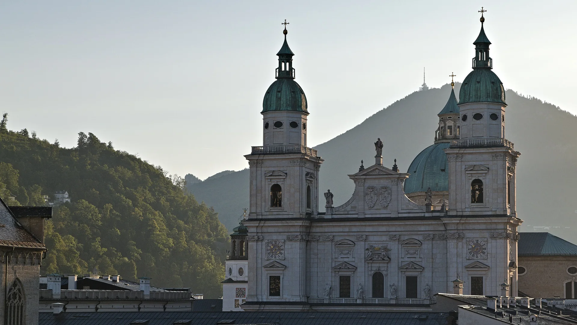 The two towers with green copper caps and the pale Baroque front of Salzburg Cathedral above the roofs of the Old Town, with a wooded mountain behind in morning light