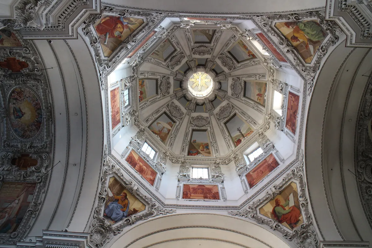Looking up into the octagonal dome of Salzburg Cathedral, with painted panels, plasterwork and a golden dove at the centre