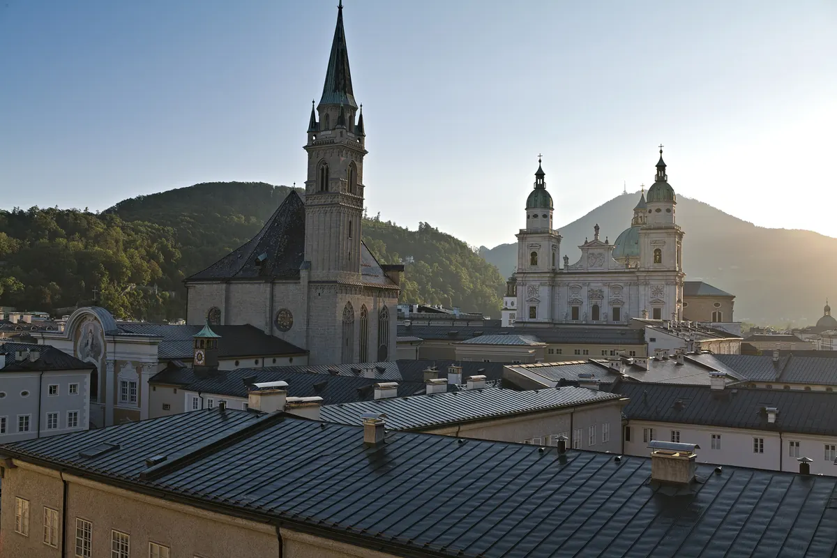The cathedral and the Franciscan church seen together above the roofs of the Old Town of Salzburg, with a wooded hill behind