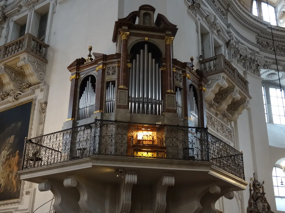 An organ case on a balcony on a pillar of Salzburg Cathedral, with gilded capitals, silver pipes and an iron balustrade in front of the keyboard